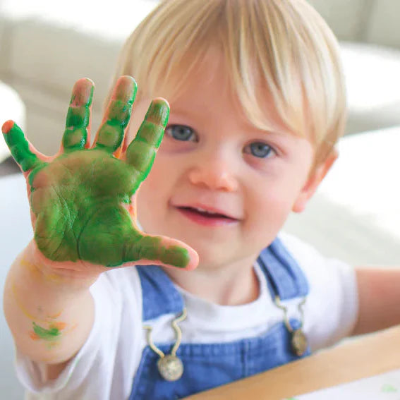 Child showing a hand with green paint, sitting at a table.