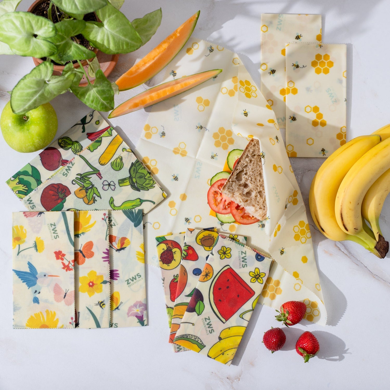 Set of reusable food wraps with fruit and vegetable designs on a white surface with fruits and vegetables.
