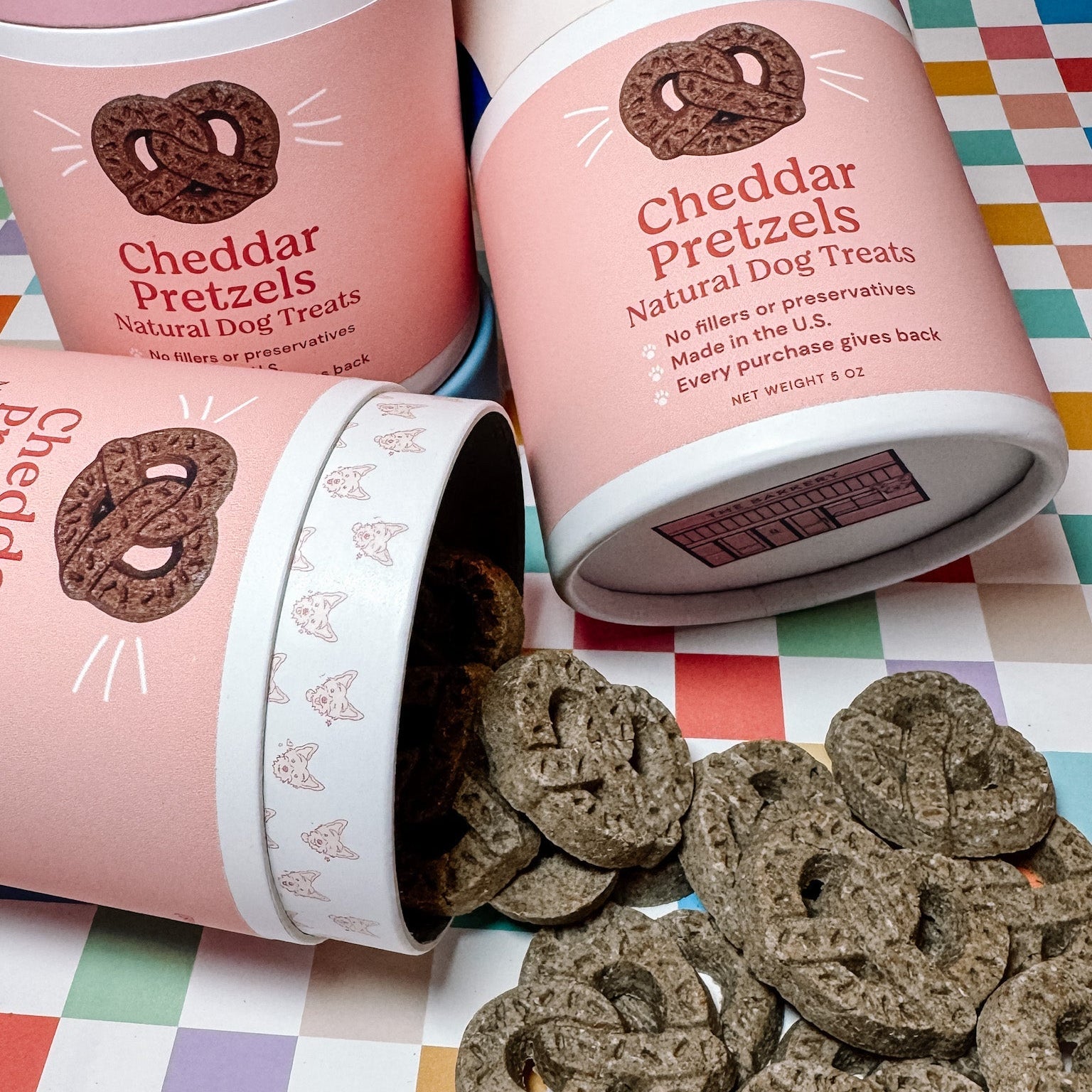 Cheddar Pretzels natural dog treats in pink containers on a colorful checkered background