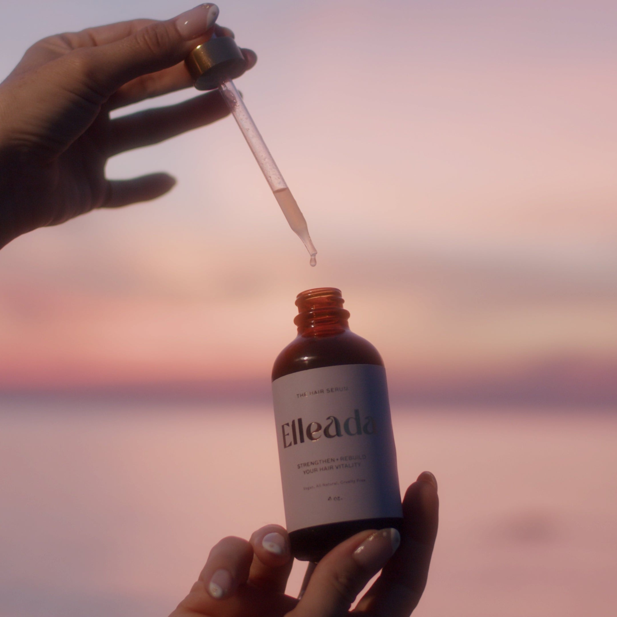 Hand holding a bottle labeled 'Elleada' with a dropper against a sunset sky.