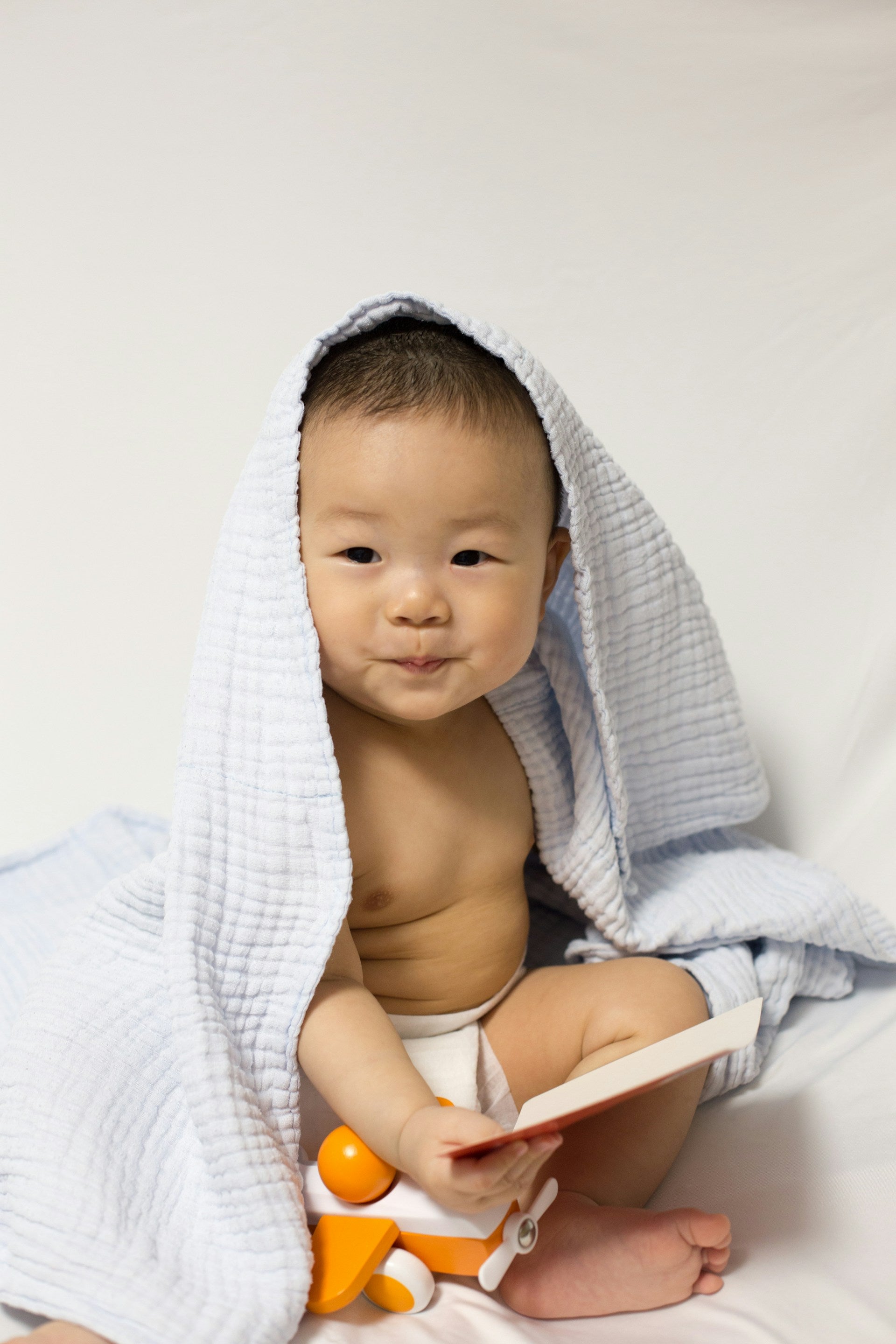 A baby boy with a blanket on his head  and a book and wooden toy on his lap.