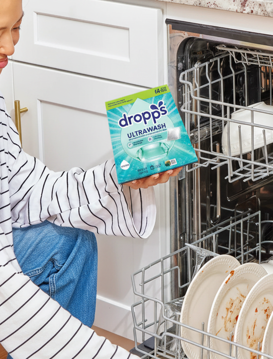 Woman in a kitchen with a dishwasher full of dishes, holding a packet of Dropps dishwasher tablets.